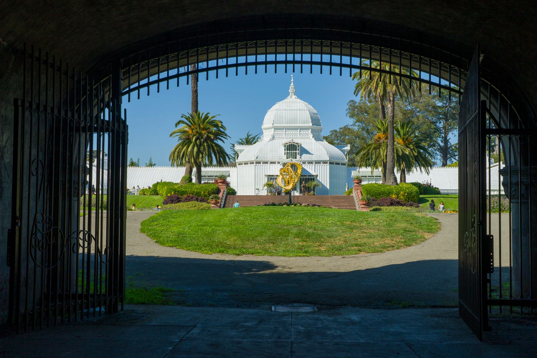 View through gates to the Conservatory of Flowers in San Francisco, featuring lush gardens and palm trees.